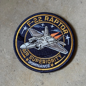 May include: A circular patch featuring an F-22 Raptor jet aircraft in shades of gray and white, with orange exhaust flames. The patch has the text "F-22 RAPTOR" and "AIR SUPERIORITY DOMINANCE" in white and yellow lettering on a navy blue background.