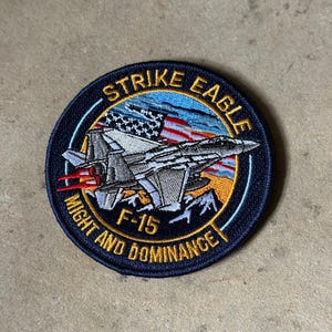 May include: A circular embroidered patch featuring an F-15 Strike Eagle fighter jet in flight with red exhaust flames. The background shows a stylized American flag and mountains. The text reads STRIKE EAGLE and MIGHT AND DOMINANCE.