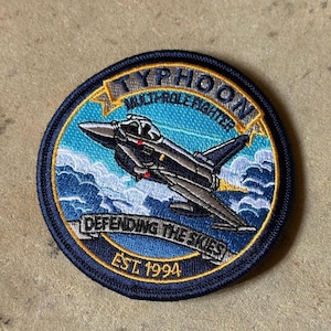 Könnte beinhalten: Runder, gestickter Aufnäher mit einem grauen und schwarzen Kampfflugzeug, der mit "TYPHOON MULTI-ROLE FIGHTER" beschriftet ist. Der Aufnäher hat einen blauen Rand mit dem Text "DEFENDING THE SKIES" und "EST. 1994". Der Hintergrund zeigt Wolken und Himmel.