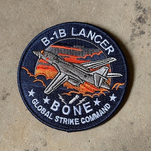 Puede incluir: Parche circular con borde azul marino que presenta un diseño de avión B-1B Lancer. El texto "B-1B LANCER" y "BONE GLOBAL STRIKE COMMAND" están bordados alrededor de la imagen del avión. El fondo muestra una puesta de sol con tonos naranja y rojo.