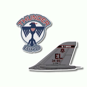 Puede incluir: Dos pegatinas con diseños de aviación. Una muestra las palabras "THUNDER BIRD" alrededor de un emblema de águila. La segunda muestra una cola de avión gris con el texto "EL" y "34 BS".