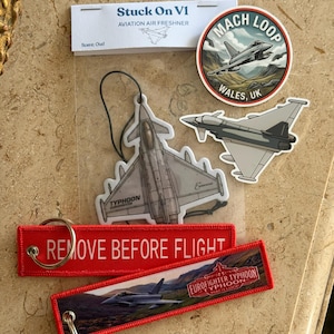 May include: A collection of aviation-themed items including a Stuck On V1 air freshener, a circular Mach Loop Wales UK sticker, a Eurofighter Typhoon sticker, and two red fabric keychains with the text Remove Before Flight and Eurofighter Typhoon.