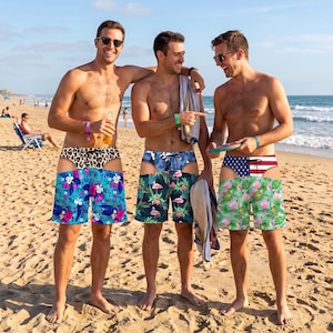 May include: Three pairs of swim trunks featuring a unique cut-out design. The first pair has a leopard print waistband and a blue floral pattern. The second pair has a blue camouflage waistband and a flamingo print. The third pair has an American flag waistband and a green tropical flamingo print.