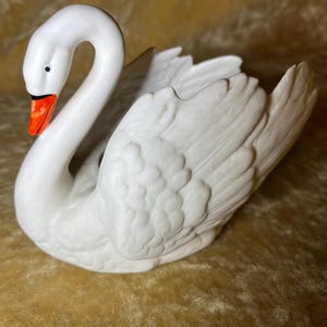 Puede incluir: Figura de cisne de cerámica blanca con cuello curvado y textura detallada de plumas. El cisne tiene un ojo negro y un pico naranja. La figura es un artículo decorativo, probablemente para exhibición.