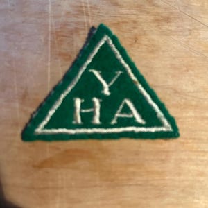 May include: A green triangular patch with white embroidered trim and the letters "YHA" in white. The patch is set against a light brown wooden surface.