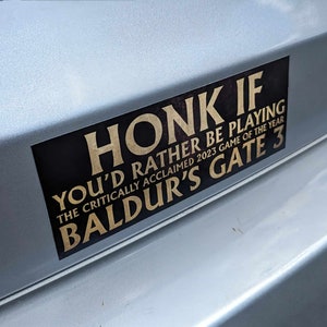 May include: A rectangular black and gold bumper sticker with the text "HONK IF YOU'D RATHER BE PLAYING BALDUR'S GATE 3 THE CRITICALLY ACCLAIMED 2023 GAME OF THE YEAR". The sticker is affixed to a silver vehicle.