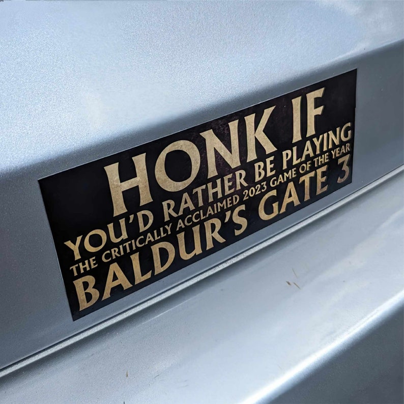 May include: A rectangular black and gold bumper sticker with the text "HONK IF YOU'D RATHER BE PLAYING BALDUR'S GATE 3 THE CRITICALLY ACCLAIMED 2023 GAME OF THE YEAR". The sticker is affixed to a silver vehicle.