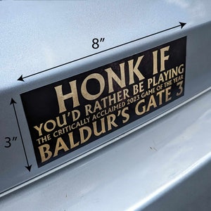 May include: A black rectangular bumper sticker, 8 inches long and 3 inches high, with gold lettering. The text reads "HONK IF YOU'D RATHER BE PLAYING THE CRITICALLY ACCLAIMED 2023 GAME OF THE YEAR BALDUR'S GATE 3."