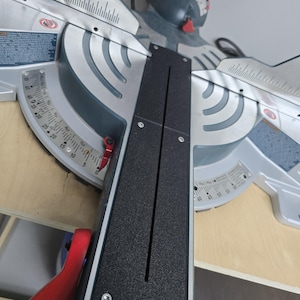 May include: A silver and dark gray miter saw featuring a black rectangular insert plate with a central slot. The tool includes a metal base with engraved angle degree markings ranging from 0 to 50 degrees, red adjustment levers, and visible screws.