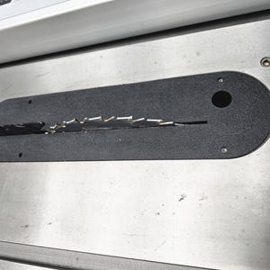 May include: A black zero-clearance table saw insert plate with a central slot for a circular saw blade. The insert is oval-shaped with a small circular hole on the right side, secured by screws into a metallic silver table saw surface.