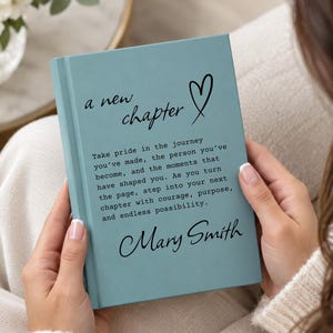 May include: A light blue hardcover journal featuring the black text "a new chapter" with a heart icon, followed by an inspirational quote about personal growth and the name "Mary Smith" printed in a cursive font on the front cover.