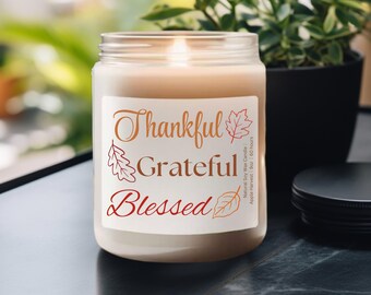 May include: A lit, scented candle in a clear glass jar. The label reads "Thankful, Grateful, Blessed" with leaf illustrations. The candle is labeled "Natural Soy Wax Candle" and "Apple Harvest Scent". The candle is 9 oz and burns for 60 hours.