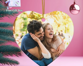 May include: Heart-shaped ceramic ornament with a photo of a smiling couple. The ornament hangs from a gold string and has a pink background. A small gift box with a ribbon is visible in the upper left corner.