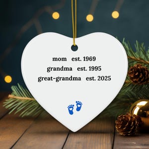 Puede incluir: Adorno de cer&aacute;mica blanco en forma de coraz&oacute;n con huellas de pies de beb&eacute; azules y el texto "mom est. 1969 grandma est. 1995 great-grandma est. 2025"