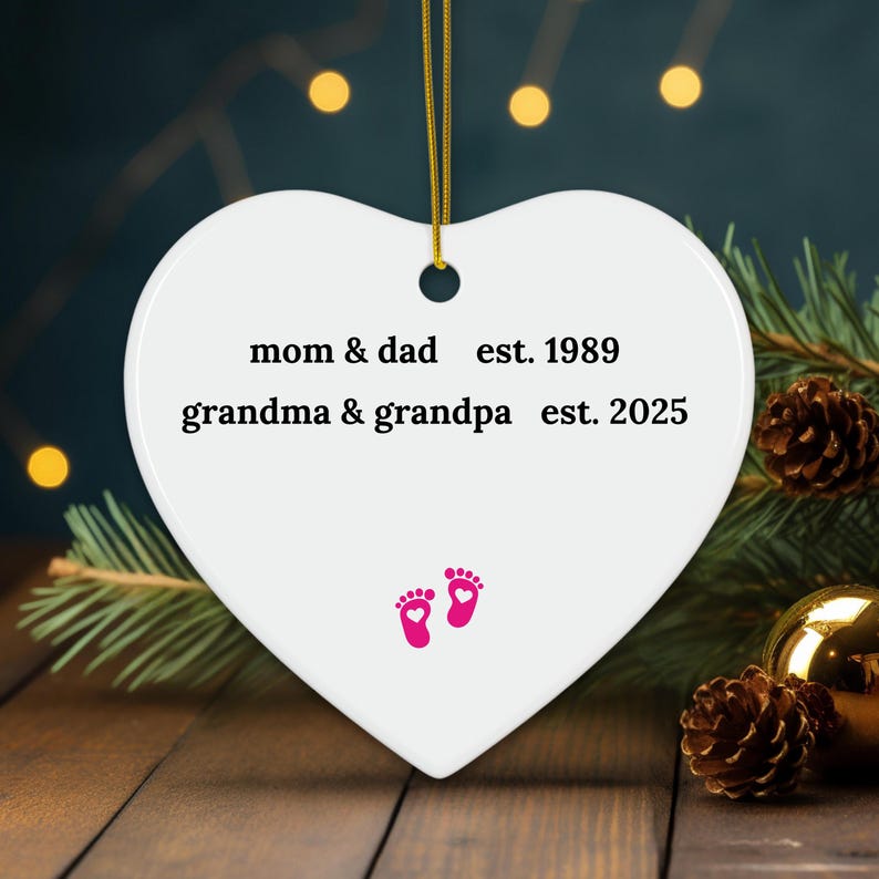 Puede incluir: Adorno de cer&aacute;mica blanco en forma de coraz&oacute;n con el texto "mom & dad est. 1989 grandma & grandpa est. 2025" y dos huellas de pies rosas con corazones.