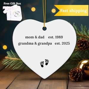 Puede incluir: Adorno de cer&aacute;mica blanco en forma de coraz&oacute;n con texto negro que dice "mom & dad est. 1989 grandma & grandpa est. 2025" y dos huellas negras con un peque&ntilde;o coraz&oacute;n en cada una.