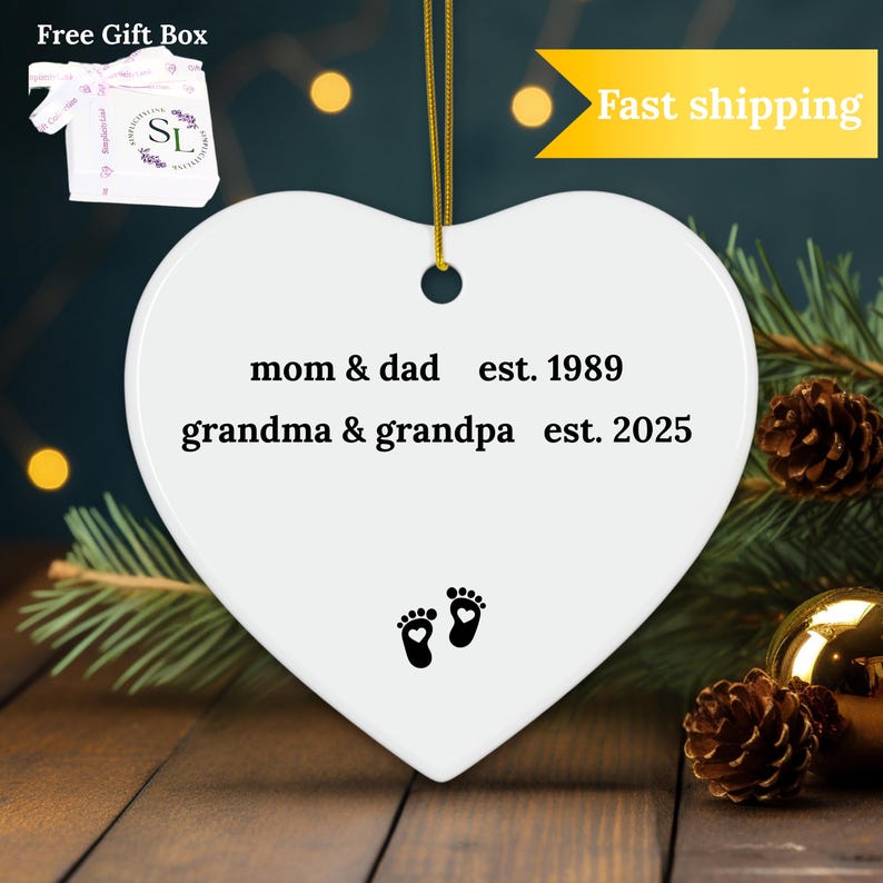Puede incluir: Adorno de cer&aacute;mica blanco en forma de coraz&oacute;n con texto negro que dice "mom & dad est. 1989 grandma & grandpa est. 2025" y dos huellas negras con un peque&ntilde;o coraz&oacute;n en cada una.