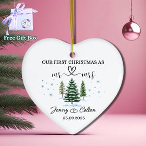 May include: A white heart-shaped Christmas ornament with the text "OUR FIRST CHRISTMAS AS mr & mrs" and the names "Jenny & Colton" with the date "05.09.2025". The ornament features illustrations of pine trees and snowflakes. A gift box with a ribbon is in the background.