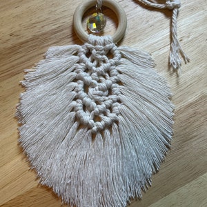 Puede incluir: Una pluma de macramé blanca con un anillo de madera y un dije de cristal. La pluma cuelga de un cordón blanco.