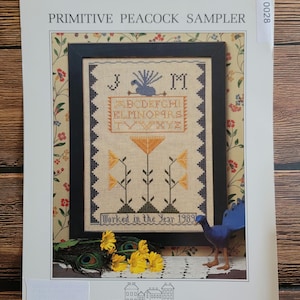 May include: A framed cross-stitch sampler titled "Primitive Peacock Sampler" with a peacock and floral design. The sampler includes the alphabet and the text "Worked in the Year 1989". The image also features a peacock figurine, peacock feathers, and yellow flowers.