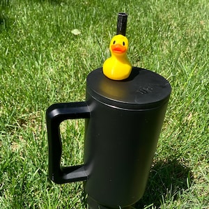 May include: A black travel mug with a yellow rubber ducky on top. The mug has a black lid and a black handle.