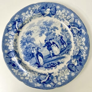 Peut inclure: Une assiette en céramique bleue et blanche avec une scène pastorale détaillée au centre. L'assiette présente un bord festonné et des motifs floraux et architecturaux sur le bord. L'image centrale représente des personnages en costumes d'époque.