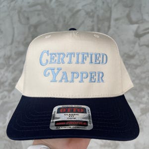 May include: A beige baseball cap with a navy blue brim. The front of the cap has the text "CERTIFIED YAPPER" embroidered in light blue. The back of the cap has a black and white label with the text "OTTO" in red, "CLASSIC FIT OSFM" in black, and "MID PROFILE MAXIMUM COMFORT" in black.