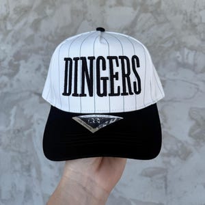 May include: A white and black baseball cap with the word "DINGERS" in large, black, block letters. The top of the cap has vertical pinstripes. The brim is solid black. A small black button is on top.