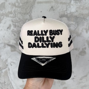 May include: A trucker hat with a black brim and a cream-colored front panel. The text "REALLY BUSY DILLY DALLYING" is embroidered in black. The hat has black stripes on the sides.