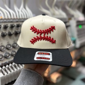 May include: A baseball cap with a cream-colored crown and a black brim. The cap features a red embroidered design resembling baseball stitching. The cap has a classic fit and is made of premium fabric.