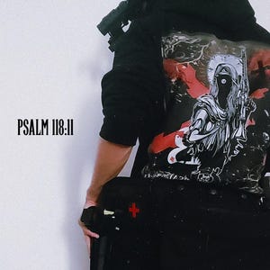 Puede incluir: Sudadera con capucha negra con un diseño gráfico en la espalda que presenta una figura sosteniendo un arma, con detalles en rojo. El texto "BLACKSPADES" está en la parte superior y "PSALM 118:11" a la izquierda. También se ve una bolsa táctica negra con una cruz roja.