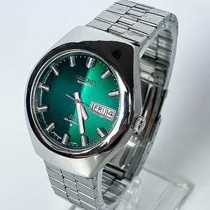 May include: A silver-tone Seiko automatic wristwatch with a vibrant emerald green sunburst dial. The watch features a stainless steel link bracelet, silver baton indices, luminous hands, and a day-date window at the three o'clock position.