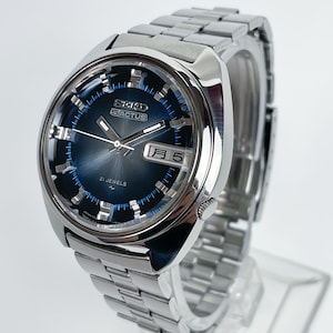 May include: A silver-toned Seiko 5 Actus wristwatch with a round, dark blue sunburst dial. The watch features a stainless steel link bracelet, silver hour markers, silver hands, and a day-date window displaying Japanese characters and the number 5.