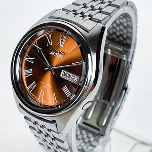 May include: A silver-tone stainless steel wristwatch with a sunburst brown dial, featuring Roman numeral hour markers, silver hands, and a day-date window at the three o'clock position. The dial displays the text Seiko Actus and 21 Jewels.