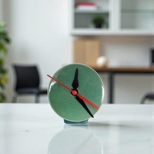 May include: A small, round, green ceramic clock with black hands and a red second hand. The clock sits on a white surface and has a light blue base. The clock's face has a textured appearance.