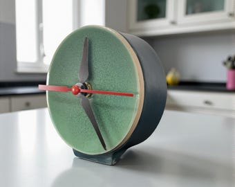 May include: A modern, ceramic clock with a teal face and black hands. The clock has a red second hand and a dark blue body. The clock is sitting on a white surface in a kitchen setting.