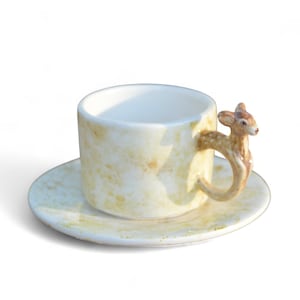 May include: A ceramic teacup and saucer set. The cup is cylindrical with a deer-shaped handle. The cup and saucer have a speckled, off-white and yellow finish. The set is designed for serving tea or coffee.