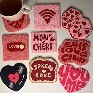 May include: A collection of handmade, textured coasters in various shapes and colors. Designs include hearts, WiFi symbols, and phrases like "LOVE," "MON CHÉRI," and "SELF LOVE CLUB." The coasters are primarily pink and red, with a black vinyl record-shaped coaster.