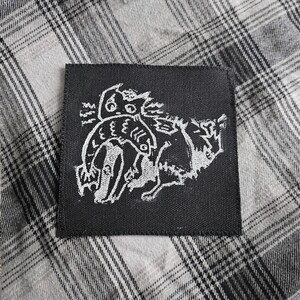 May include: Square black patch with a white illustration of a creature with multiple eyes and a long tail. The patch is on a gray and white plaid fabric.