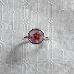 May include: A silver ring featuring a circular bezel setting that contains a small, flat inlay with a pink and white floral pattern. The flower design displays delicate pink petals radiating from a textured, brownish-yellow centre point.