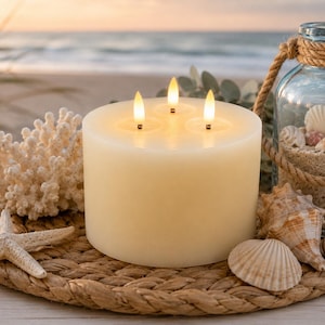 May include: A cylindrical off-white candle with three lit LED flames sits on a round woven rope mat. The arrangement includes a white coral piece, a starfish, various seashells, and a clear glass jar filled with sand and shells, tied with rope.