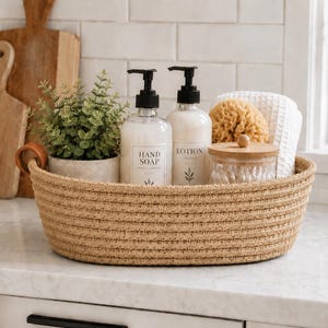 May include: An oval woven rope basket with a tan finish and a small brown leather handle. Inside are two clear pump bottles labeled Hand Soap and Lotion, a small potted green plant, a round glass jar with a wooden lid, and a white textured cloth.