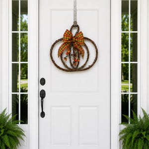May include: A fall-themed pumpkin wreath made of woven twigs, adorned with a plaid bow in shades of orange, green, and yellow. The wreath is decorated with small, berry-like ornaments. It hangs on a white door.