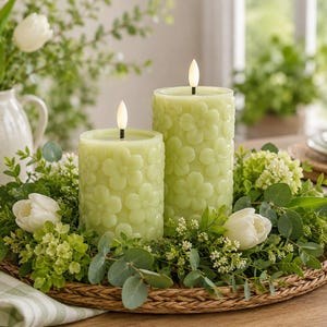 May include: Two light green cylindrical LED candles with a raised floral pattern on the exterior, each topped with a small white flame-shaped bulb, arranged on a round woven tray surrounded by white tulips and green foliage.