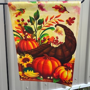 May include: A fall-themed garden flag featuring a cornucopia overflowing with pumpkins, sunflowers, and berries. The flag has a yellow background with colorful autumn leaves and is hanging from a black metal hook.