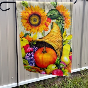 May include: A decorative garden flag featuring a cornucopia overflowing with autumn harvest items. The design includes a large orange pumpkin, grapes, pears, apples, and sunflowers against a wooden background. The flag is hanging on a black metal stand.