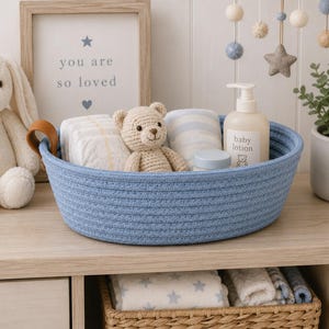 May include: A round blue woven rope basket with a tan leather handle containing a small crocheted teddy bear, folded white diapers, a striped cloth, and a white plastic bottle labeled baby lotion with a small bear graphic on the front.