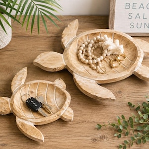 May include: Two light brown wooden trays shaped like sea turtles with a carved shell pattern. The larger tray holds a string of wooden beads and gold rings, while the smaller tray contains a black car key fob and a silver metal key.
