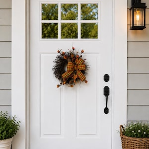 May include: A white front door with a wreath, a black handle, and a window with a view of trees. The wreath has a plaid bow and fall-themed accents. A black lantern and potted plants are also visible.
