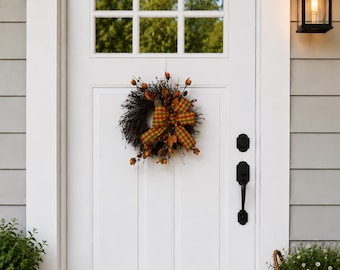 May include: A white front door with a wreath, a black handle, and a window with a view of trees. The wreath has a plaid bow and fall-themed accents. A black lantern and potted plants are also visible.